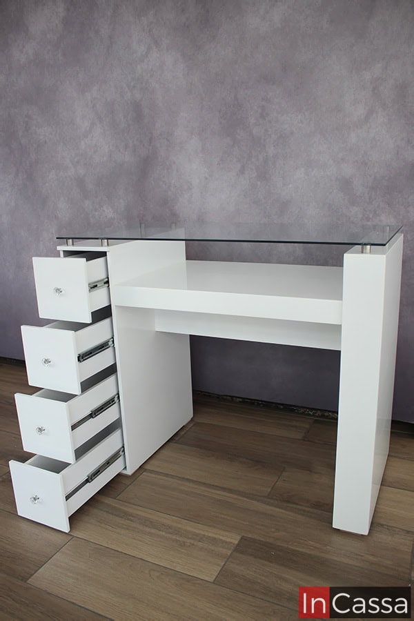 La toma nos deja ver la mesa de uñas desde una vista lateral que nos permite conocer más a detalle su estructura, mostrando el grosor de sus patas, la estructura completa de sus cajones y algunos detalles adicionales como son los separadores metálicos de la cubierta de cristal. Adicionalmente, destaca el color blanco de la pintura presenta en la totalidad de la mesa.
