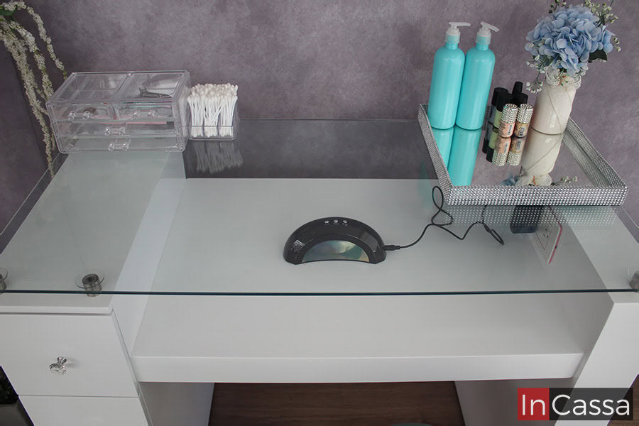 La foto se concentra en la cubierta de cristal, dejándonos observar sus separadores metálicos que le proporcionan una posición suspendida en la mesa, además del espacio para instalar lampara de uñas el cual cuenta con un contacto eléctrico. Todo el mueble es presentado en un atractivo color blanco brillante.