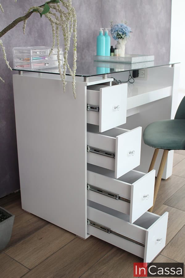 Vemos la mesa de uñas desde una toma lateral de la pata con cajones, los cuales se encuentran abiertos mostrando su estructura, misma que cuenta con rieles metálicos incorporados para su sistema de apertura y cierre. Al igual que todas las partes de la mesa, los cajones mantienen el color blanco en todas sus partes.
