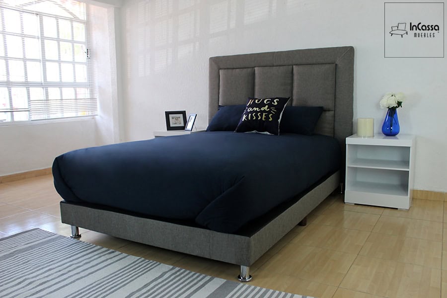 Una toma lateral resalta la elegante recámara tapizada en lino mouse. Destacan la cabecera acolchada en bloques simétricos, patas metálicas cromadas y dos mesas de noche blancas, creando un espacio moderno y luminoso.