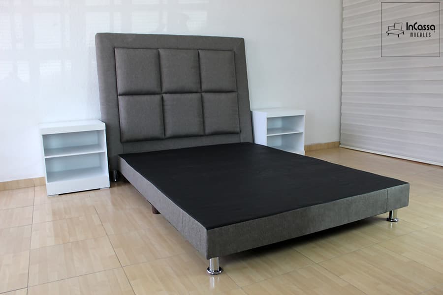 Una toma en ángulo derecho muestra la estructura completa de la base tapizada en lino mouse, acompañada de una cabecera con diseño acolchado en cuadros y burós blancos de líneas limpias. Las patas metálicas elevan el conjunto con un toque moderno.