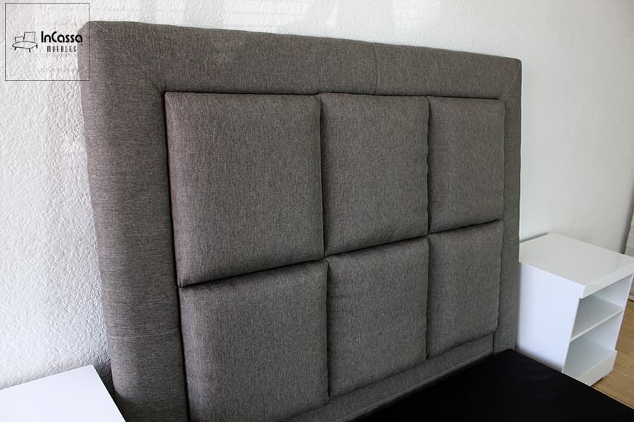 Una toma en ángulo izquierdo destaca la textura de la cabecera tapizada en lino mouse, con diseño acolchado en cuadros simétricos. El contraste con los burós blancos resalta el estilo moderno y elegante de la recámara.