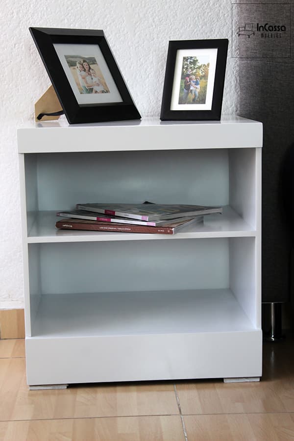 La imagen muestra un buró blanco con acabado brillante, capturado de frente. Sus dos repisas abiertas ofrecen espacio funcional para libros o revistas, mientras que la parte superior se decora con marcos fotográficos que aportan un toque personal.