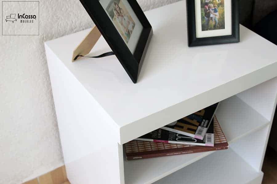 Una toma en ángulo superior destaca el acabado brillante del buró blanco, resaltando su superficie lisa y esquinas definidas. Sobre él, dos portarretratos decoran con calidez, mientras que las repisas inferiores albergan revistas de forma ordenada.