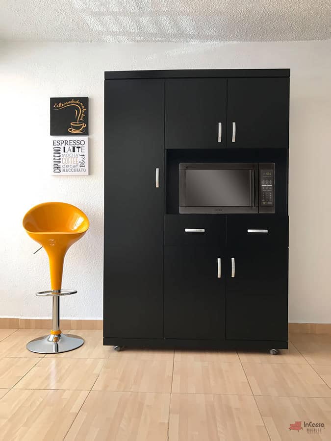 La imagen muestra la elegante Alacena para Microondas MÁLAGA en acabado chocolate mate, visto de frente y en plano completo. Destaca su diseño funcional con múltiples compartimentos y espacio central para microondas, adornado con jaladeras metálicas plateadas. A la izquierda, un banco alto amarillo crea un contraste vibrante.