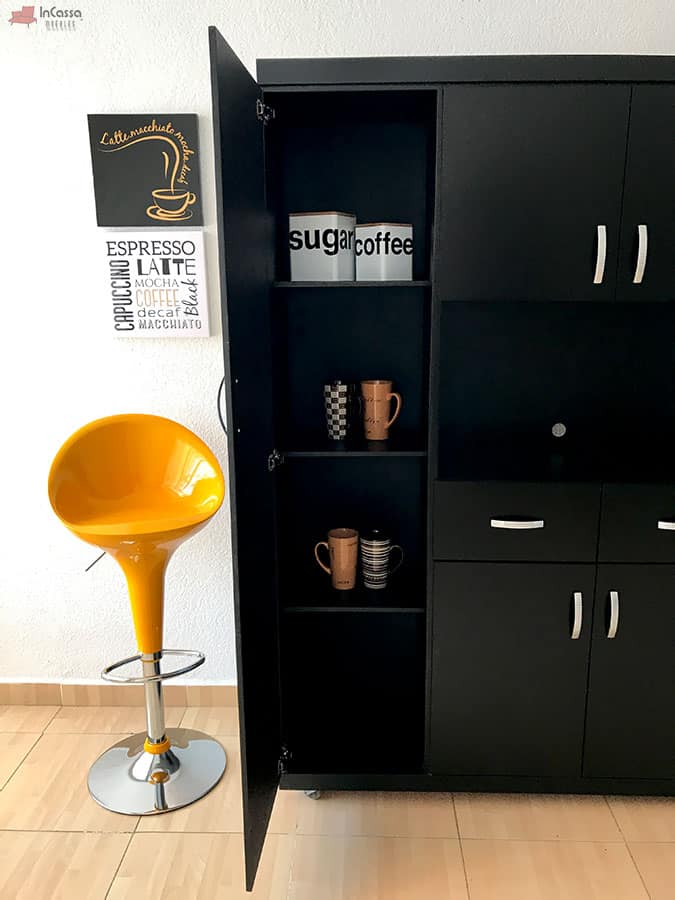 La foto presenta la Alacena para Microondas MÁLAGA en acabado chocolate mate con la puerta lateral izquierda abierta, mostrando tres repisas internas organizadas con frascos de azúcar y café, así como tazas decorativas. El contraste lo aporta un banco alto amarillo brillante junto a la pieza.