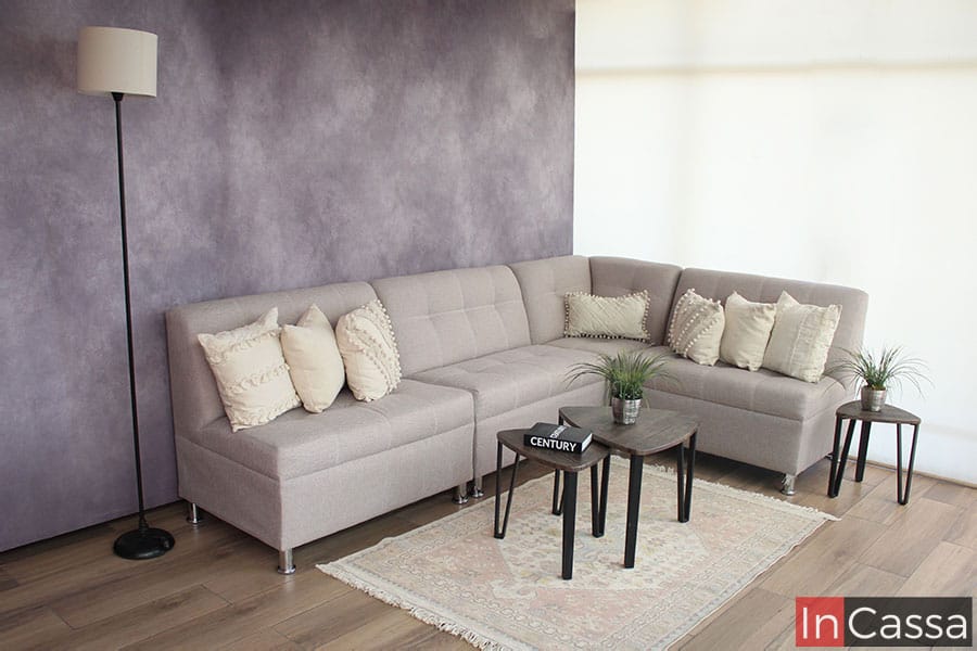 Sala en escuadra tapizada en tela lino color greige, sostenida por pequeñas patas metálicas en los extremos del esquinero y los 3 love seat que conforman el mueble. La sala se encuentra instalada en una estancia con una pared gris y piso de madera.