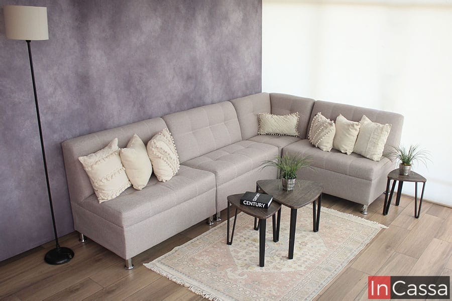 Una foto completa de la sala en escuadra tapizada totalmente en lino color greige, adornada con algunos artículos decorativos (no incluidos en el precio) tales como unas mesitas de centro y cojines pequeños. El mueble se presenta esquinada junto a una pared gris y una ventana completa, además de un piso de madera.