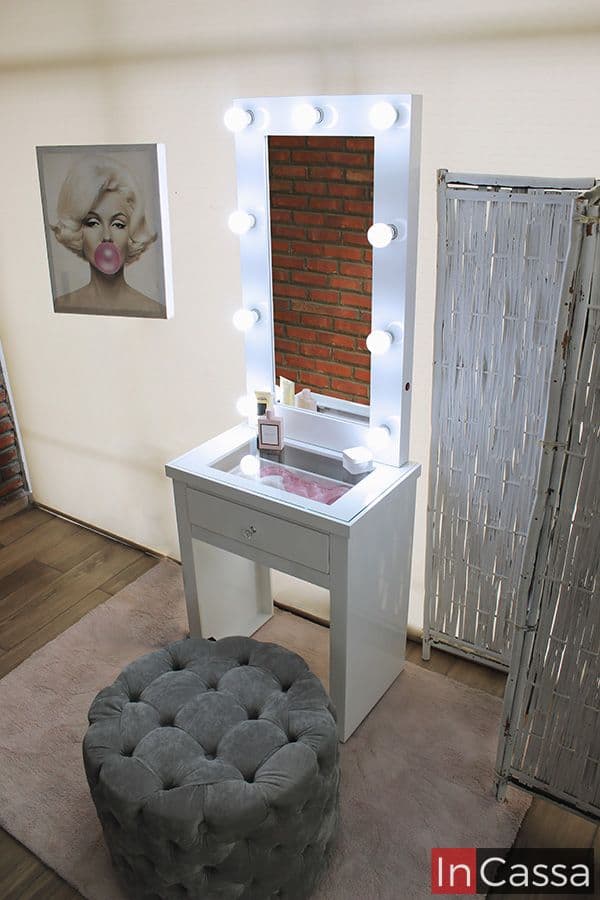 La imagen presenta un tocador blanco ideal para espacios reducidos, con un diseño moderno y funcional. Incluye un espejo vertical rodeado por 9 focos LED de luz blanca tipo Hollywood, un cajón inferior central y superficie con vidrio transparente que permite visualizar el contenido del compartimento superior.