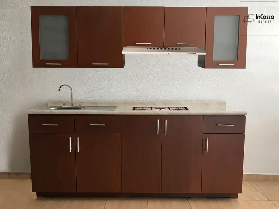 La imagen muestra una cocina integral en elegante tono maple oscuro, compuesta por módulos superiores con puertas lisas y vitrinas, y módulos inferiores con cubierta de formica color claro. Incluye espacio para parrilla y tarja de acero inoxidable.