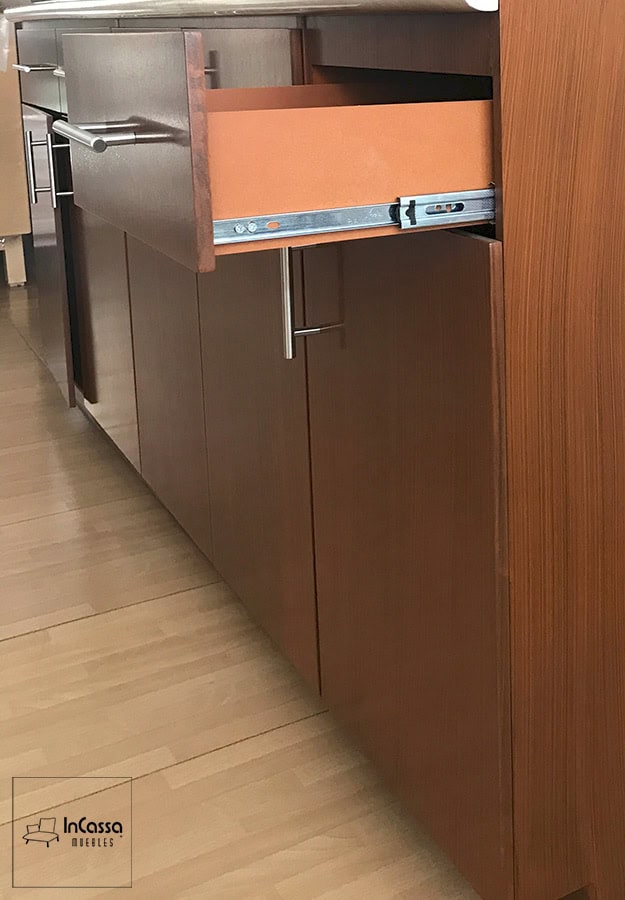 Una toma lateral muestra el detalle de un cajón abierto con corredera metálica de extensión y acabado interior en tono maple. La cocina tiene acabado en color maple oscuro y puertas con jaladeras metálicas.