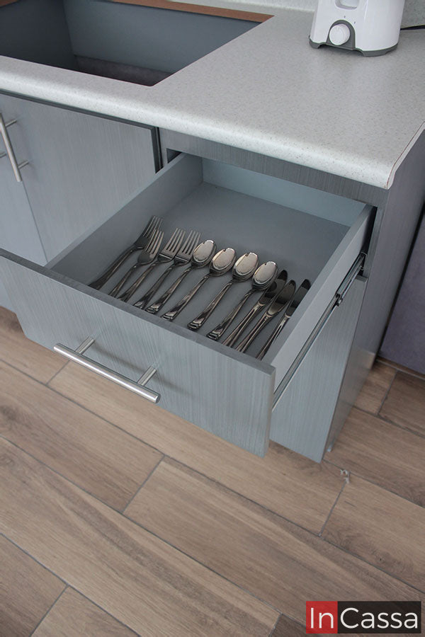 La imagen presenta un cajón superior extraído, con divisiones internas que almacenan cubiertos, en una cocina con acabado en gris cenizo claro. El frente del cajón incluye jaladeras metálicas modernas que combinan funcionalidad y diseño elegante.
