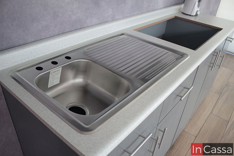 Una toma desde arriba muestra la tarja de acero inoxidable con escurridor integrado, perfectamente montada sobre una cubierta de formaica estilo sal y pimienta. Los gabinetes inferiores, en tono gris cenizo claro, complementan el estilo moderno y funcional de la cocina.