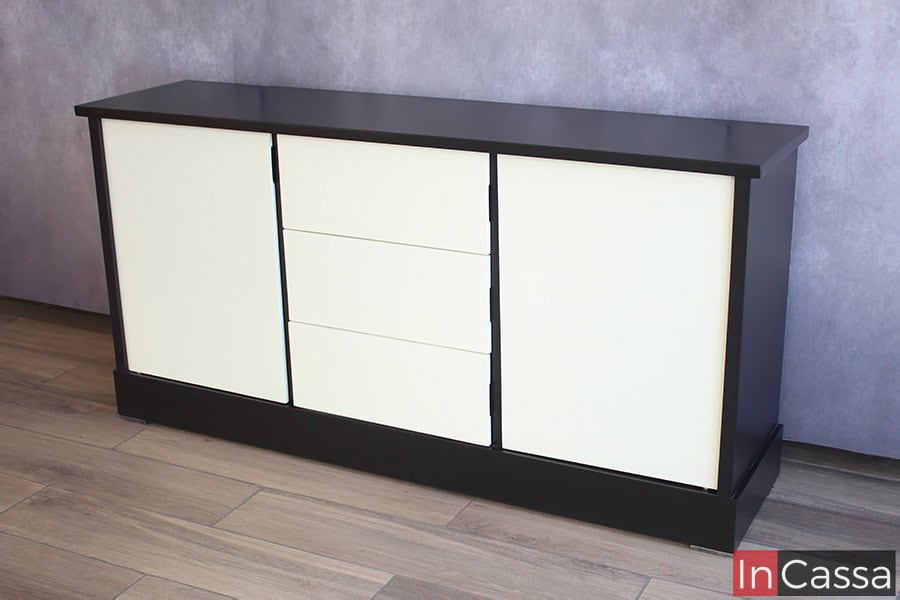 Se presenta el bufetero desde una toma lateral, en la que se puede apreciar de manera más completa la estructura del mueble, la cuál es de color negro y cuenta con cajones y puertas de color blanco.