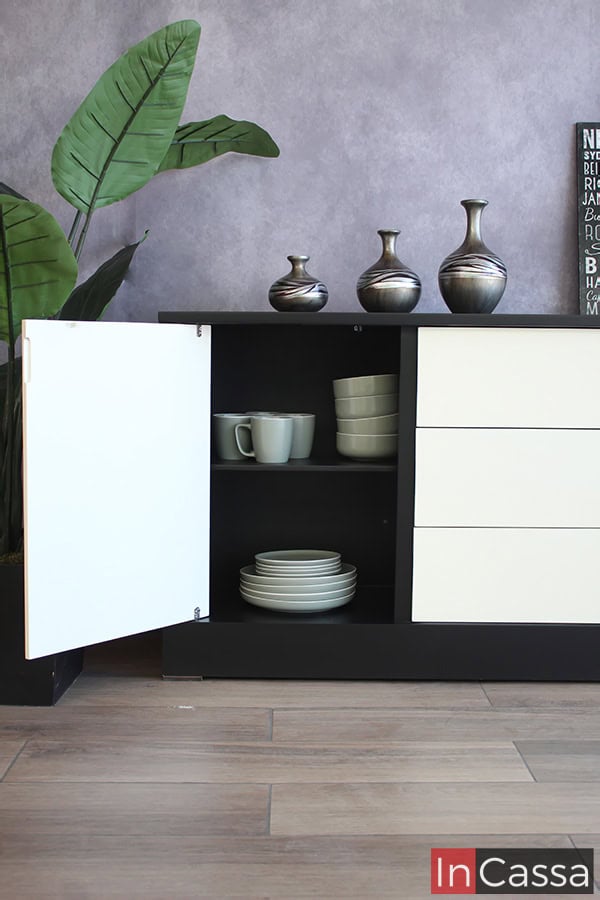 Una foto enfocada en el espacio lateral izquierdo del bufetero (viéndolo de frente), resaltando su interior gracias a que se muestra con su puerta blanca abierta. Es sección cuenta con dos compartimentos divididos por un entrepaño, adornados por unas cuantas vajillas que además ayudan a determinar la amplitud de cada espacio, cabe mencionar que el interior mantiene el mismo color negro que el resto de la estructura del bufetero.