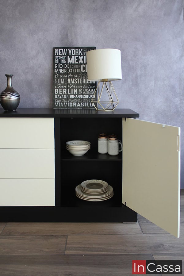 La imagen se concentra en mostrar los compartimentos con los que cuenta el espacio lateral derecho del bufetero (desde una vista frontal frontal), en el que podemos ver un entrepaño que divide el espacio en dos compartimentos para guardar vajillas y utensilios. El interior de esta sección es de color negro al igual que las partes exteriores, y además cuenta con una puerta blanca completa.