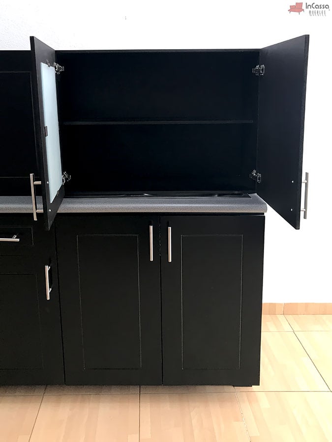 La imagen muestra una amplia alacena superior de doble puerta, una de ellas con acabado en vidrio esmerilado, que ofrece gran capacidad de almacenamiento con entrepaño interno. Justo debajo se encuentra el módulo inferior con tarja de acero inoxidable integrada en la cubierta de formaica estilo granito purpina, complementado con puertas y jaladeras metálicas modernas. Ambos módulos en acabado chocolate completo.