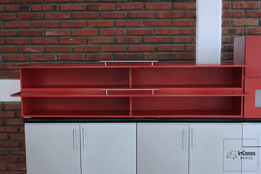 Una fotografía de las alacenas superiores del modelo MEDELLÍN con las puertas abatibles abiertas, revelando amplios compartimentos dobles con repisas internas. El contraste del rojo vivo contra el muro de ladrillo aporta un estilo moderno e industrial.