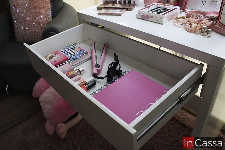 La imagen muestra el cajón abierto del tocador blanco, mostrando una cuidada organización con libretas, productos de belleza, una plancha de cabello rosa y accesorios. Sobre el tocador, se observan más artículos de maquillaje y joyería, todo en un ambiente ordenado y elegante.