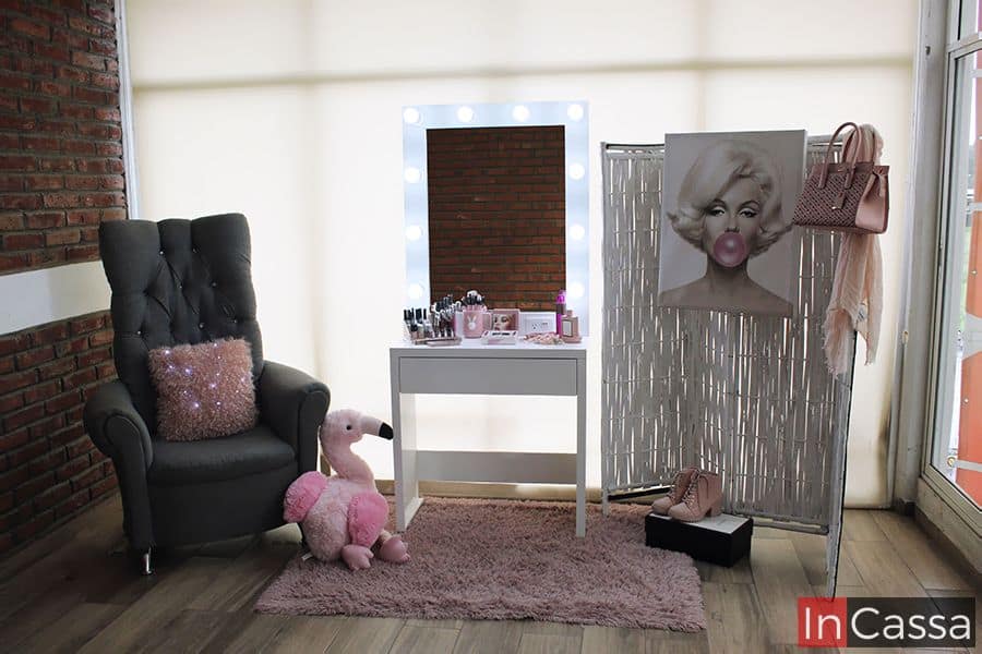 Tocador blanco con un espejo iluminado por luces LED, decorado con artículos de maquillaje y perfume, sobre una alfombra rosa. Al lado, un elegante sillón gris capitoné y un divisor de ambiente con pintura de Marilyn Monroe.