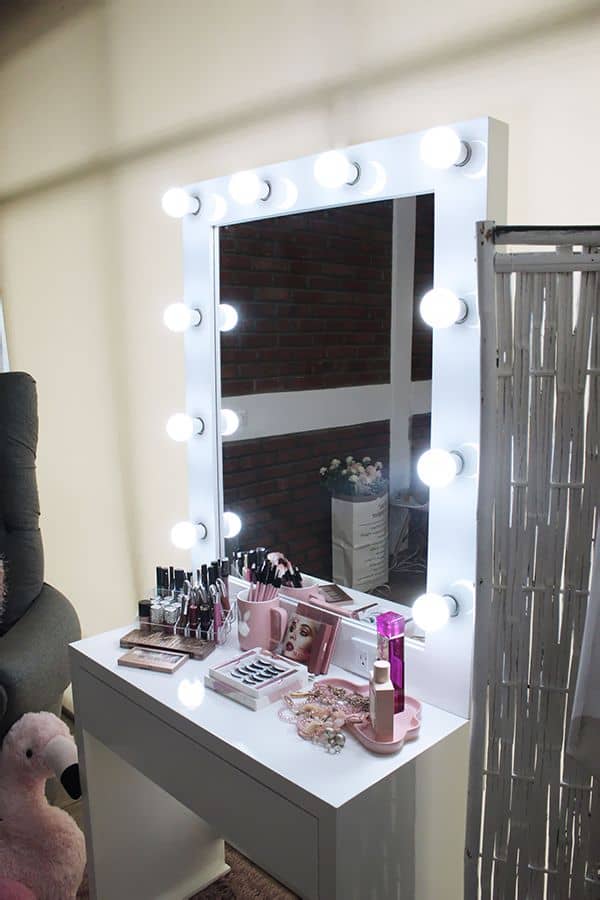 Toma lateral del tocador blanco con un espejo de luces LED brillantes. Sobre la superficie, una variedad de productos de maquillaje, brochas, labiales y accesorios. El ambiente se complementa con un cómodo sillón gris y un divisor decorativo al fondo.