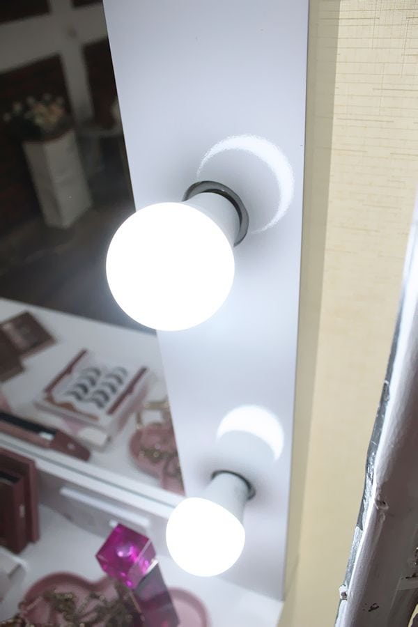 Primer plano de las brillantes luces LED del espejo de un tocador blanco. En el reflejo, se observan productos de maquillaje y accesorios sobre la superficie del tocador, creando un ambiente glamoroso y ordenado.