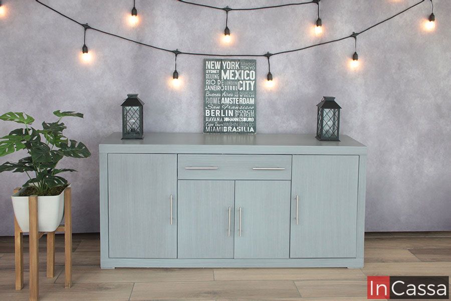 La imagen presenta el Bufetero Vintage Modelo MISSISSIPPI en un elegante tono gris cenizo claro, capturado de frente para resaltar su composición simétrica y líneas rectas. Se aprecian sus tres puertas amplias y dos cajones superiores con jaladeras metálicas alargadas, así como su acabado liso y moderno. Al fondo, un ambiente decorativo con luces cálidas y elementos minimalistas complementa perfectamente el estilo del mueble.