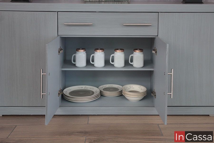 Una toma frontal revela el compartimento central del Bufetero MISSISSIPPI en gris cenizo claro, mostrando su amplitud y organización. Se aprecian dos niveles: arriba, jarras decorativas bien acomodadas; abajo, platos y tazones apilados. Las puertas abiertas lucen bisagras metálicas resistentes y jaladeras modernas que complementan su estilo vintage–minimalista.