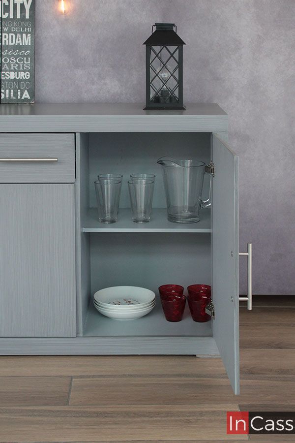 La imagen muestra el costado derecho del Bufetero MISSISSIPPI en gris cenizo claro, destacando su interior amplio y bien distribuido. En la repisa superior se aprecian vasos y una jarra transparente, mientras que en la repisa inferior se acomodan platos y vasos decorativos rojos. La puerta abierta revela bisagras metálicas resistentes y jaladera moderna que realzan su estilo vintage–minimalista.