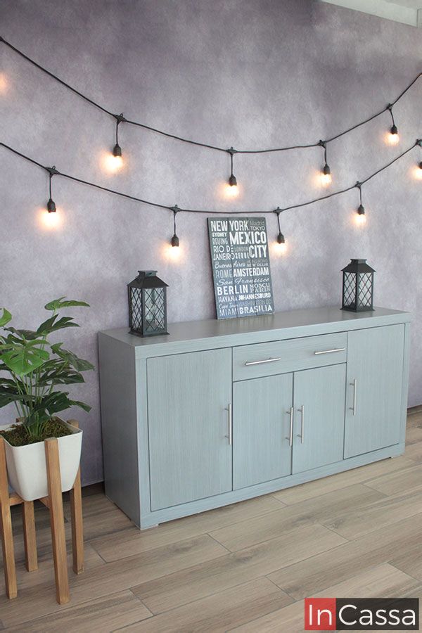 La imagen muestra una toma lateral del Bufetero Vintage Modelo MISSISSIPPI en color gris cenizo claro, destacando su acabado homogéneo y textura suave. El ángulo resalta la profundidad del mueble, sus puertas amplias con jaladeras metálicas y los cajones superiores perfectamente alineados. La escena decorada con luces cálidas, una planta y faroles negros crea un ambiente acogedor que realza su estilo moderno y elegante.
