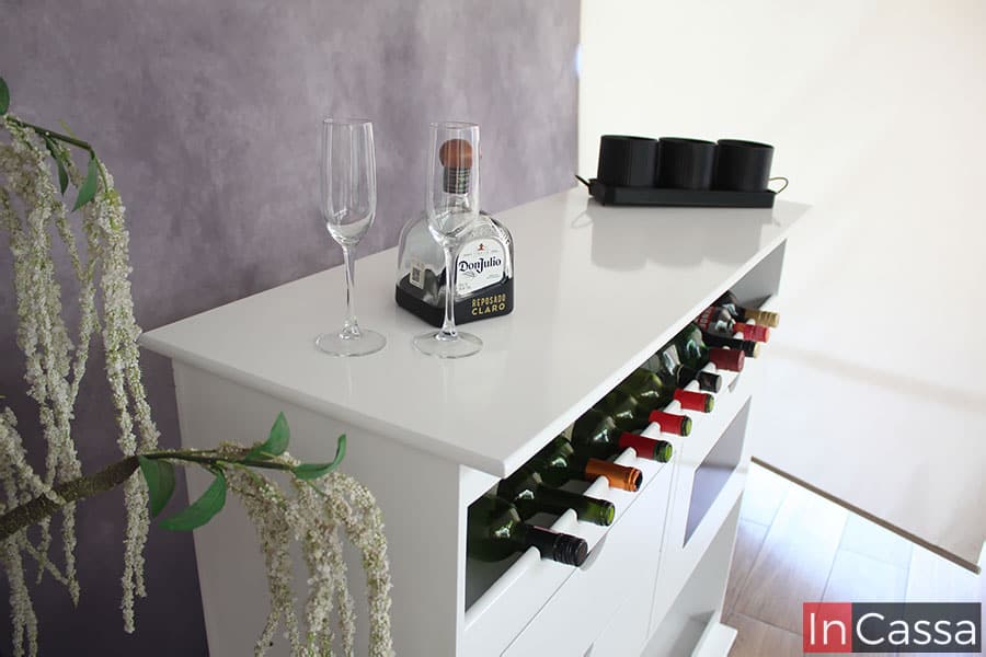 Mejor vista de la cubierta y la parte lateral de la cantina; como podemos ver, cada parte del mueble está pintado en color blanco con acabado brillante, mismo que queda mucho más resaltado en la cubierta de MDF Chileno gracias a las copas y botella que la adornan y se reflejan en el acabado de la pintura. Además también se puede apreciar la cava y algunos de los compartimentos de la cantina.