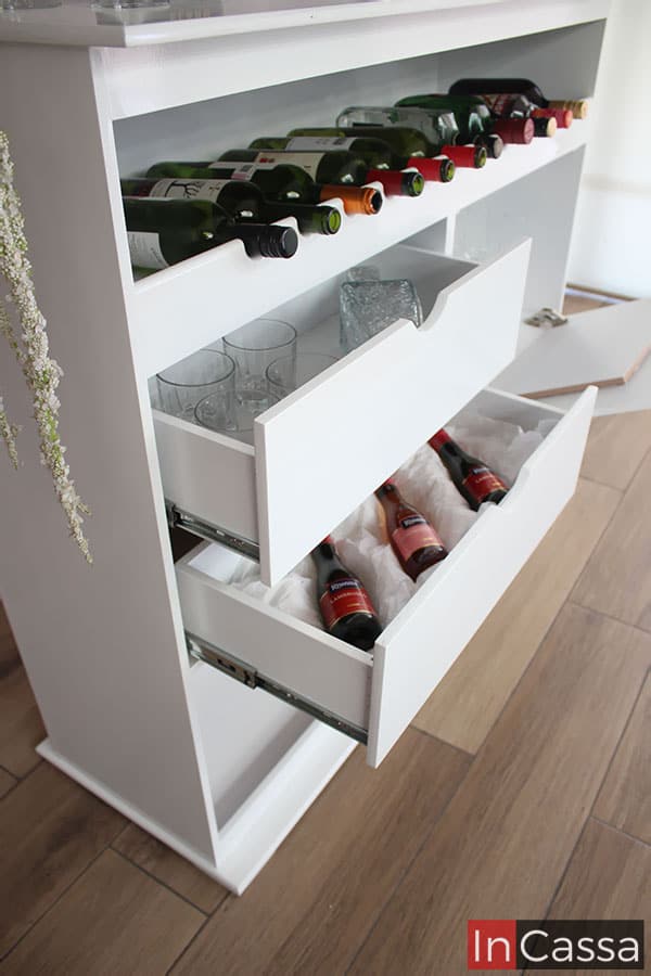 Una foto más enfocada en mostrarnos la cava incorporada en la estructura de la cantina, en la cuál podemos observar 11 espacios para guardar botellas. Además también se nos muestra la construcción de los cajones, misma que es completamente blanca al igual que todo el mueble, cada cajón cuenta con un sistema de apertura y cierre guiado por rieles metálicos, sin mencionar que se decora su interior con algunos vasos de cristal y botellas a fin de ejemplificar la capacidad de cada uno.