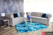 La imagen muestra una sala con un sofá y un love seat estilo capitoné tapizados en suede gris perla, decorados con cojines azules. La alfombra azul vibrante y las mesas modernas crean un ambiente elegante y acogedor.