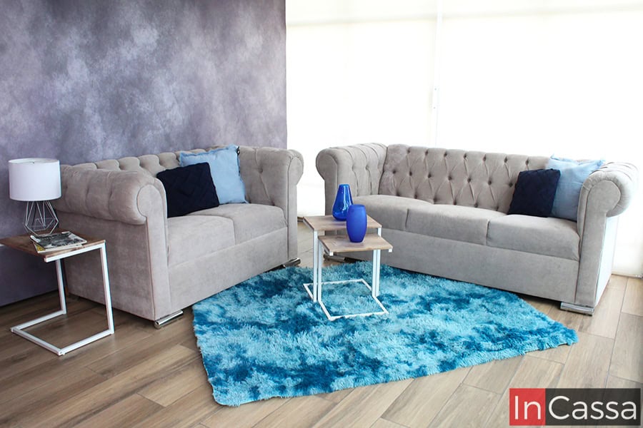La imagen muestra una sala con un sofá y un love seat estilo capitoné tapizados en suede gris perla, decorados con cojines azules. La alfombra azul vibrante y las mesas modernas crean un ambiente elegante y acogedor.