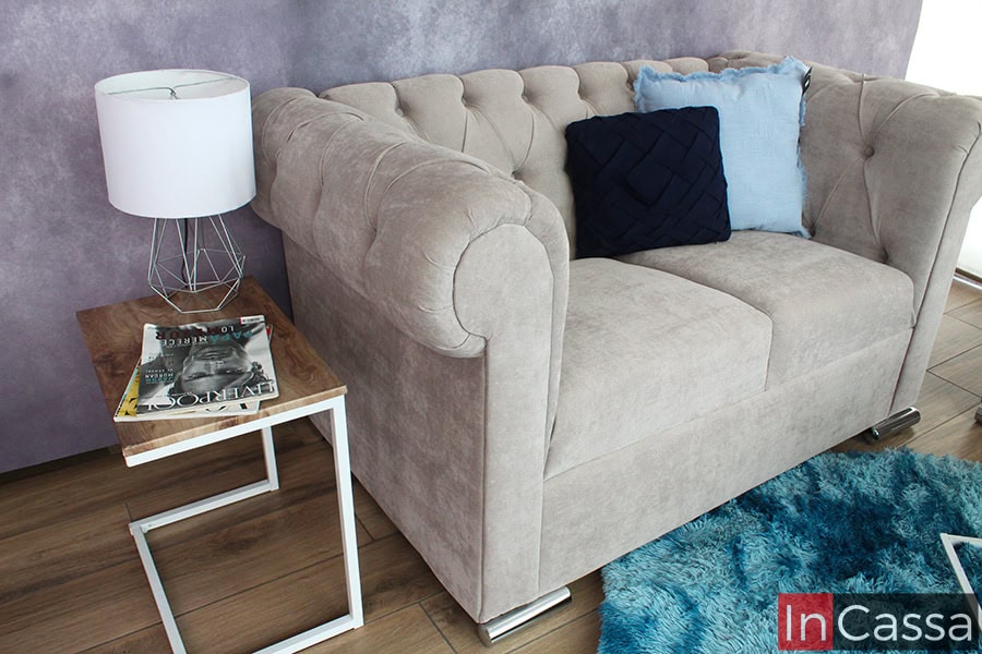 La foto presenta el love seat estilo capitoné en suede gris perla, acompañado de cojines azul oscuro y celeste. A su lado, una mesa auxiliar moderna sostiene una lámpara decorativa y una revista, complementando el ambiente sofisticado.