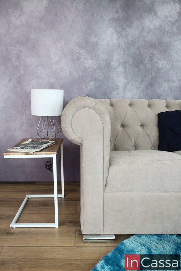 Una toma cercana del love seat capitoné tapizado en suede gris perla, mostrando los detalles curvos de los brazos y el diseño acolchado. Junto a él, una moderna mesa lateral con una lámpara y revistas, aportando un toque elegante al espacio.