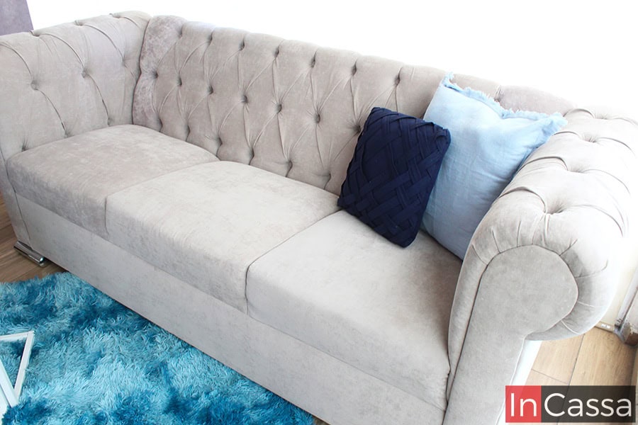 La imagen muestra un sofá capitoné de tres plazas en suede gris perla, decorado con cojines azul oscuro y celeste. El diseño acolchado del respaldo resalta la elegancia clásica, mientras que la alfombra azul añade un toque moderno y vibrante.