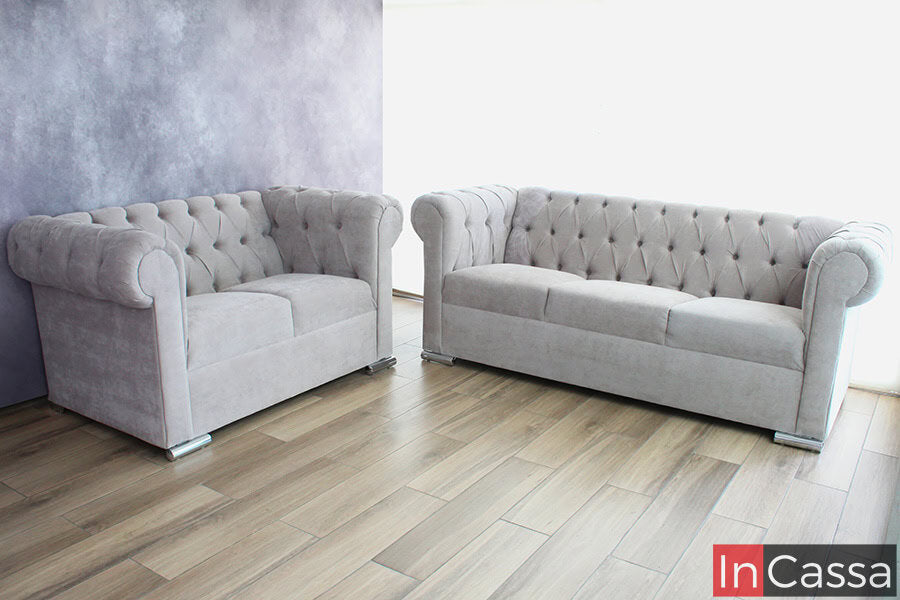 La imagen presenta un conjunto de sofá y love seat en estilo capitoneado en suede gris perla, con un diseño elegante y clásico. Los módulos de dos y tres plazas, combinados con patas metálicas cromadas, aportan un toque sofisticado y moderno al ambiente.