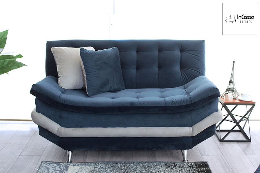 La foto presenta el love seat del modelo Murcia en un ángulo frontal, donde resalta su tapizado en suede azul marino con franja decorativa en tono hueso. Su diseño curvo y acolchado con costuras capitonadas ofrece una apariencia envolvente y elegante, acompañado por cojines decorativos que realzan su confort y estilo contemporáneo.