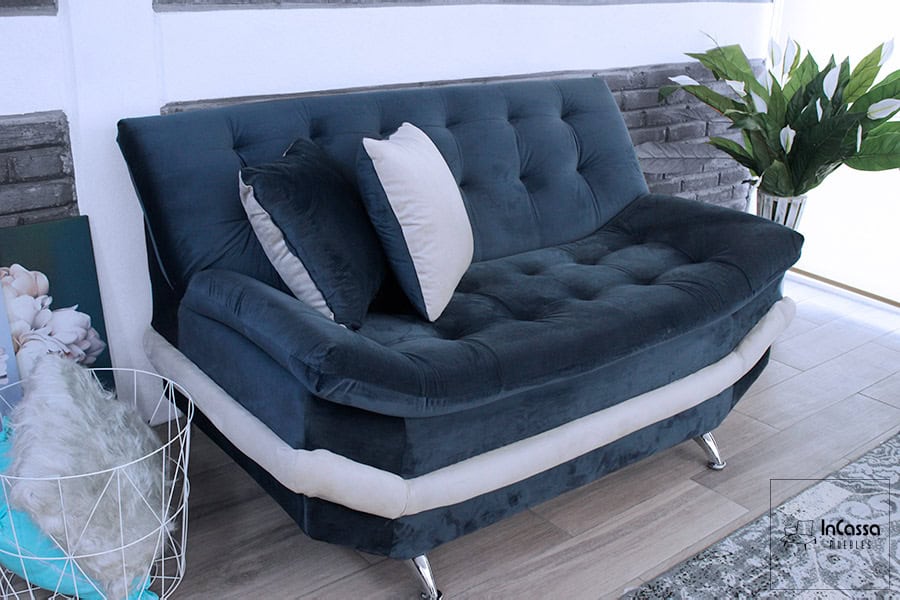 La fotografía presenta el love seat del modelo Murcia, tapizado en suede azul marino con detalles en tono hueso. Su estructura robusta y diseño capitonado destacan sobre un fondo neutro con muros de ladrillo gris y plantas naturales, acentuando su estilo moderno y su elegancia atemporal.