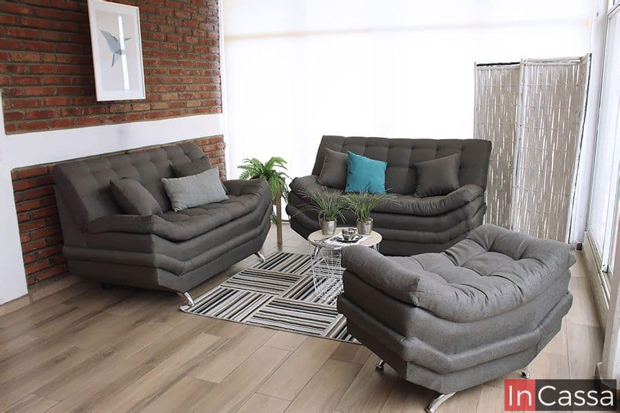 La imagen muestra la Sala Modelo Murcia en una elegante versión tapizada en lino mouse, destacando su diseño contemporáneo y sus líneas capitonadas que aportan volumen y confort. La composición con dos love seat y taburete crea un ambiente acogedor, mientras los cojines en tonos neutros y azul turquesa añaden un toque moderno ideal para salas luminosas y con estilo urbano.