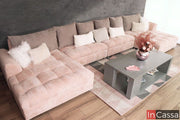 Una toma superior en ángulo muestra esta sala esquinera tapizada en suede rosa palo, con 2 chaise longue y cojines decorativos en tonos neutros. La textura aterciopelada del tapizado y la mesa central gris sobre alfombra geométrica completan un ambiente acogedor y moderno.