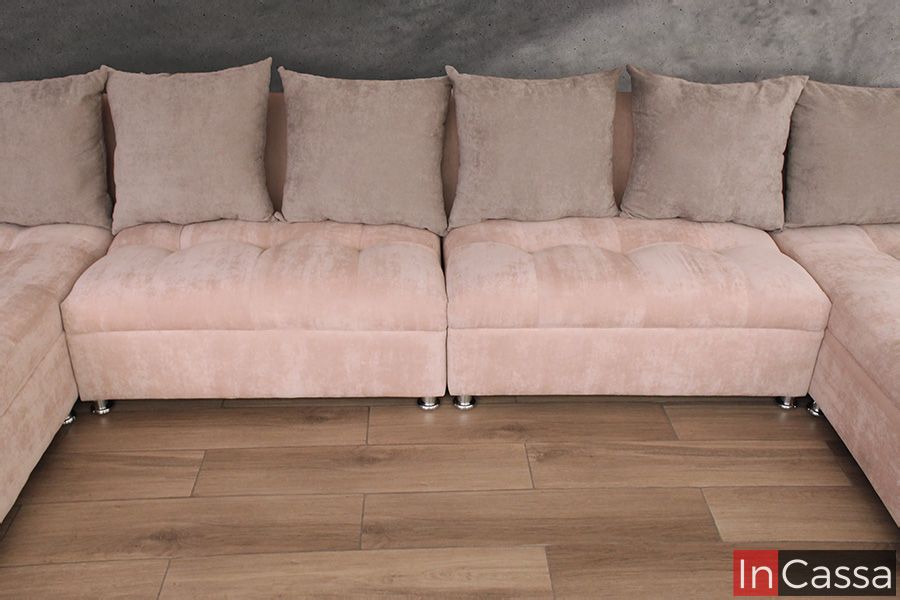 Una toma frontal resalta los asientos centrales de la sala esquinera en tapiz rosa palo. El tapizado con diseño capitonado y cojines respaldo en beige crean una combinación armoniosa sobre el piso tipo madera y fondo gris elegante.