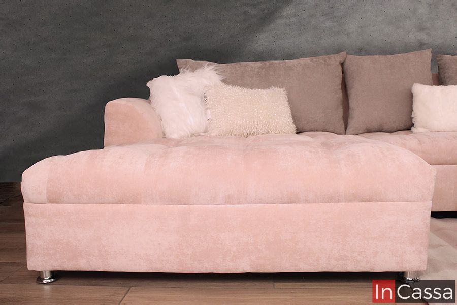 La foto muestra de frente el chaise longue en tapiz suede rosa palo aterciopelado y patas metálicas cromadas. Los cojines blancos y beige de distintas texturas aportan calidez, mientras que el fondo gris acentúa su diseño elegante.