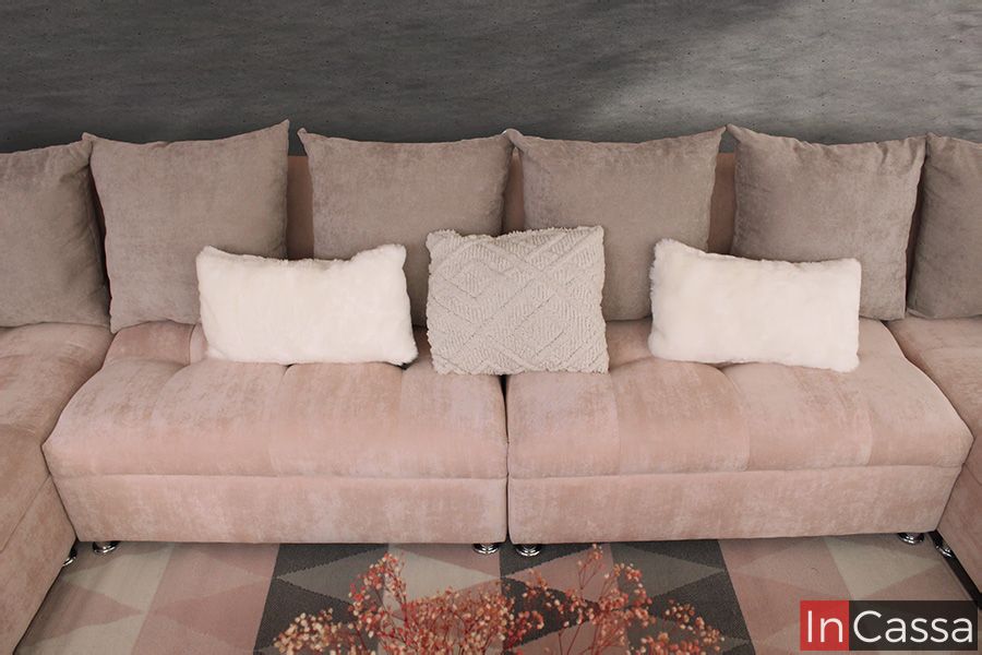 Una toma superior detalla los asientos centrales de la sala esquinera tapizados en tela suede rosa palo. Los cojines decorativos en blanco y beige, junto al patrón de la alfombra inferior, crean un ambiente armónico y lleno de confort.