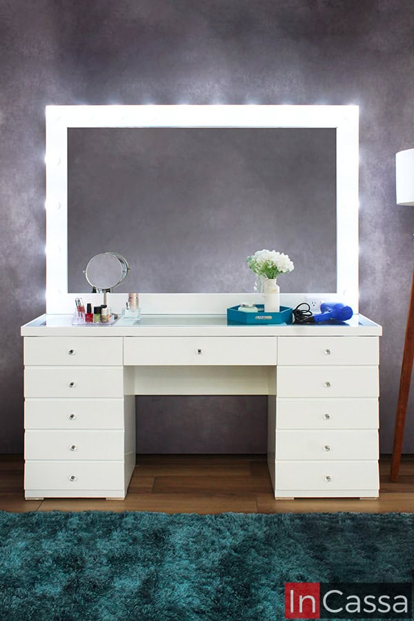 Se presenta el tocador Mykonos blanco en vista frontal, mostrando sus 11 cajones con jaladeras estilo diamante que aportan distinción. El espejo iluminado con 18 luces LED enmarca el diseño, creando un ambiente moderno, funcional y lleno de estilo para el área de maquillaje.