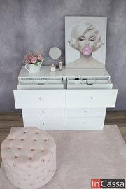 Tocador blanco de moderno diseño con ocho cajones, de los cuales, los dos superiores cuentan con dos compartimentos extras ocultos. El mueble es presentado con distintos utensilios y artículos decorativos, instalado frente a una pared gris.
