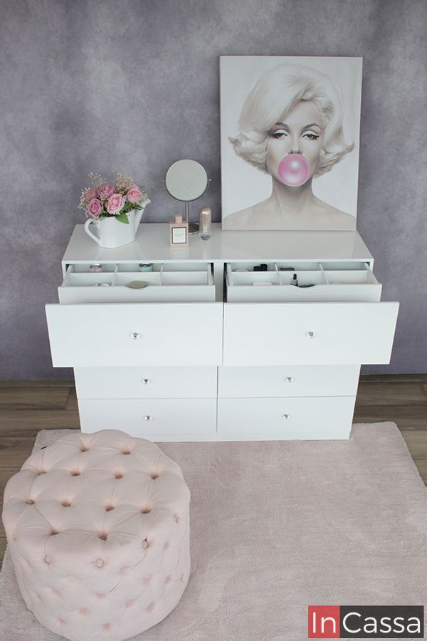 Tocador blanco de moderno diseño con ocho cajones, de los cuales, los dos superiores cuentan con dos compartimentos extras ocultos. El mueble es presentado con distintos utensilios y artículos decorativos, instalado frente a una pared gris.