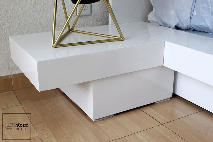 La imagen destaca el diseño geométrico y moderno del buró en acabado blanco brillante. Su forma suspendida y estructura sólida complementan perfectamente la base tipo plataforma, aportando funcionalidad con un estilo minimalista elegante.