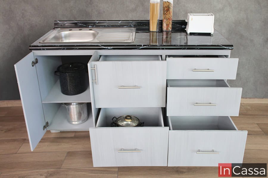 La toma se enfoca completamente en el módulo inferior largo, el cuál es presentado con sus cajones y puertas abiertos, dejándonos observar sus compartimentos internos, los cuales son de diferentes tamaños y capacidades para guardar todo tipo de utensilios. El mueble también tiene una cubierta de formaica estilo mármol negro y una tarja de acero inoxidable, además de que la totalidad de su estructura es de acabado gris cenizo claro.,